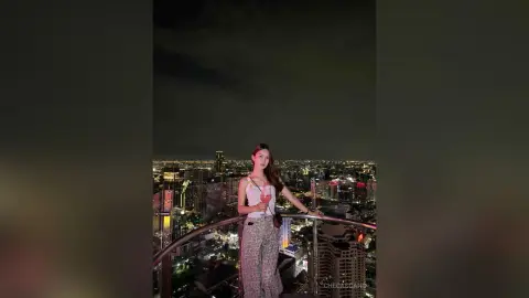 CheCascano poses on a rooftop balcony at night overlooking a bright city skyline, wearing a white tank top and patterned pants.