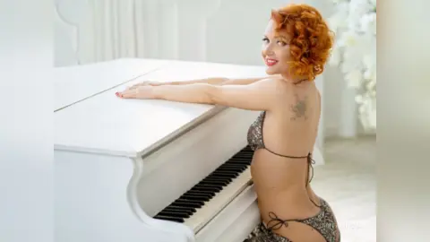Smiling redhead woman in a sparkly bikini leaning on a white grand piano in a bright, elegant room.