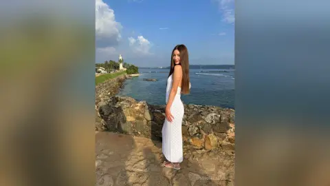 Smiling woman in a white summer dress standing by a stone wall overlooking the ocean on a sunny day.