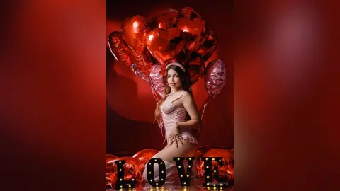 Glamorous woman in pink lingerie posing amidst red heart balloons for a romantic Valentine's Day photoshoot.