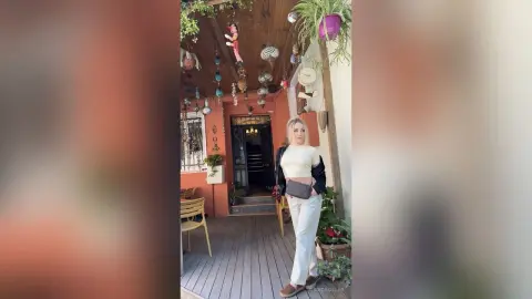 Blonde woman in white trousers and black cardigan standing on a rustic wooden patio decorated with hanging plants.