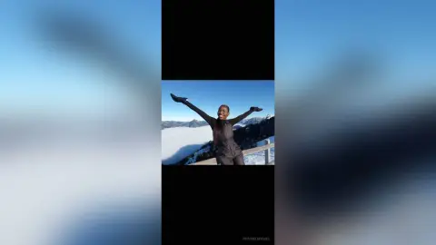 Joyful woman in black outfit raising arms against a scenic mountain lake landscape under a clear blue sky.