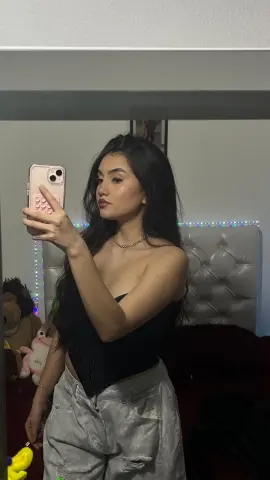 Young woman in a black corset and grey denim taking a mirror selfie in a cozy bedroom setting.