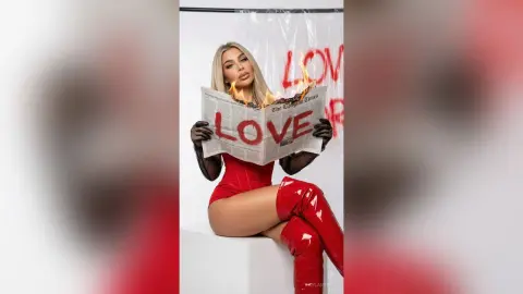 Glamorous woman in a red bodysuit and tall boots holding a newspaper with LOVE text in a studio setting.