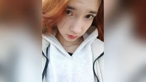 Young woman with reddish hair wearing a cozy white hoodie in a bright, casual indoor setting.