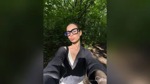 CarlynKernell wearing glasses and a black zip-up jacket while posing outdoors in a sunny, wooded area.