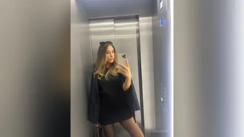 VictoriaWynn poses for a mirror selfie in an elevator, wearing a short black dress and a dark plaid jacket over her shoulders.