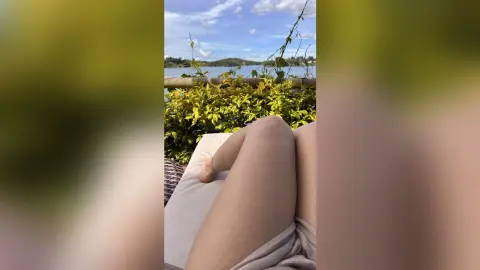 Relaxed person lounging on a patio chair overlooking a scenic lake and lush greenery on a sunny day.