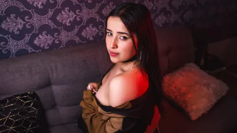 Young woman in a black off-the-shoulder top sitting on a grey sofa against patterned wallpaper in moody lighting.