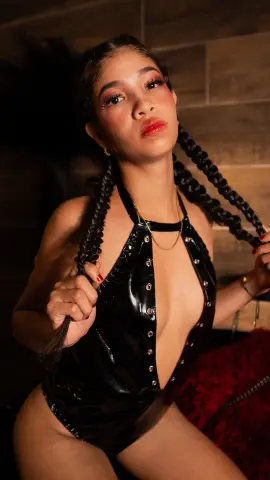 Young woman with long braids in a black latex bodysuit posing in a moody, dimly lit indoor setting.