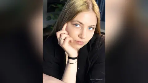 Portrait of a blonde woman in a black shirt looking thoughtfully at the camera in a soft-lit indoor setting.