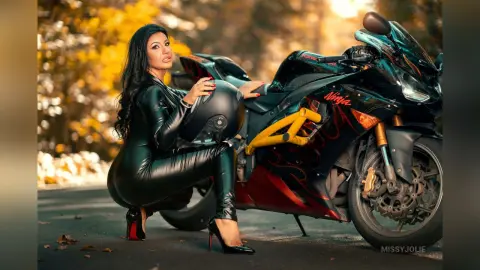 MissyJolie poses next to a black motorcycle wearing a tight black leather bodysuit and red high heels.