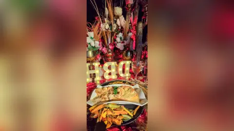 Elegant dinner table setting with gourmet food, floral centerpiece, and glowing gold letters on a red tablecloth.