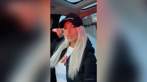 Blonde woman in a black hoodie and baseball cap sitting in a car with a relaxed mood.