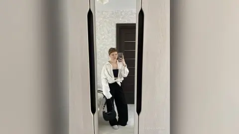 Young woman in a white button-down shirt and black trousers taking a mirror selfie in a modern minimalist room.