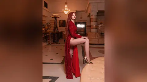 RubyAttwood poses in a long red dress with a high slit, showing off her bare leg and gold high heels in a luxury room.
