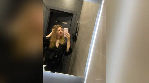 Young woman in a black outfit taking a mirror selfie in a modern minimalist bathroom.