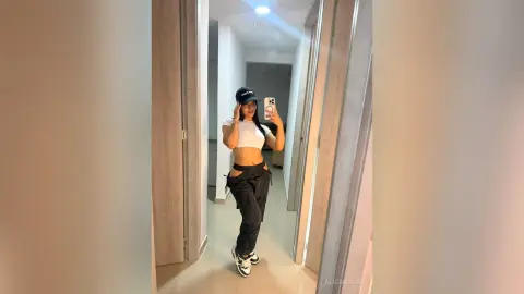 Young woman in white crop top and black cargo pants taking a mirror selfie in a modern hallway.