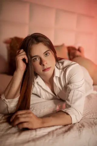 Young woman in a white button-down shirt relaxing on a bed with warm, cozy lighting.