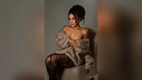 Glamorous woman in a fuzzy faux fur coat and patterned tights sitting elegantly in a studio setting.