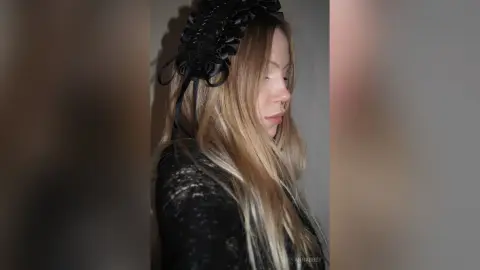 Blonde woman in a black lace outfit wearing a black floral headband in a moody indoor setting.