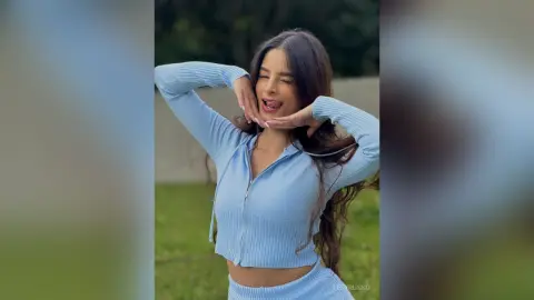 TefyRusso poses outdoors with her hands near her face, sticking her tongue out while wearing a light blue long-sleeve zip-up top.