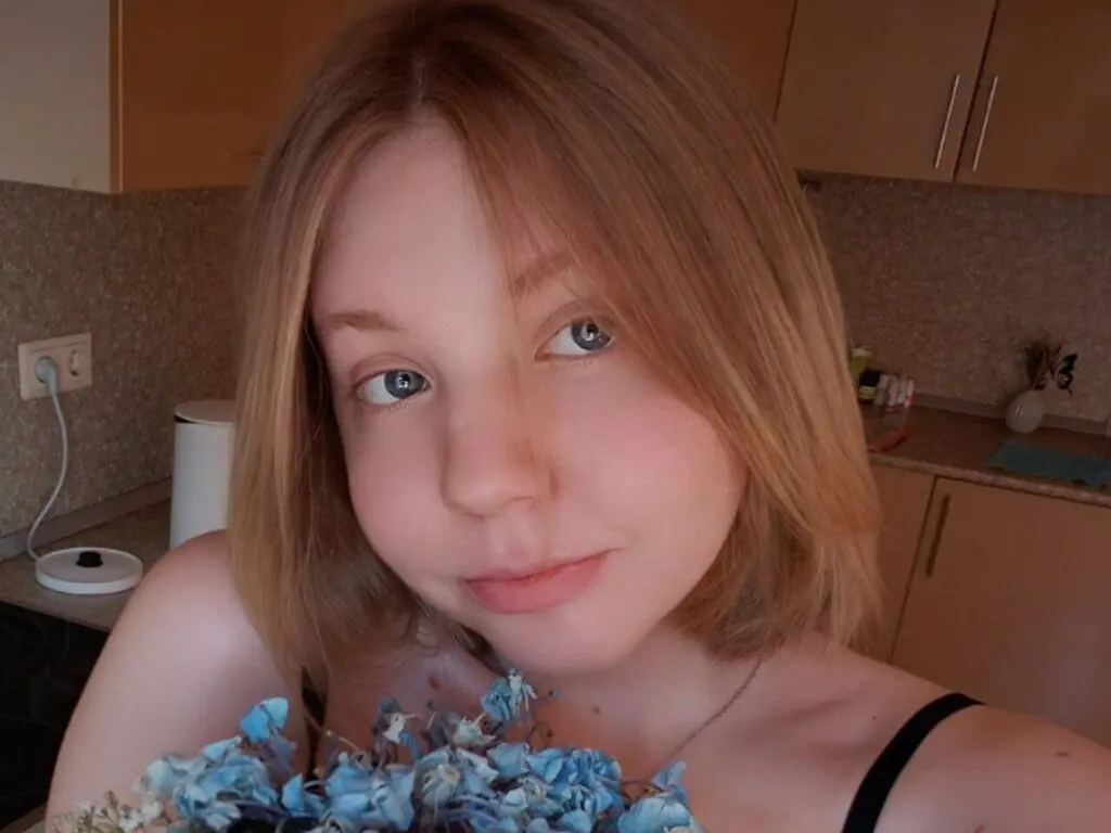 Blonde woman with blue eyes holding blue flowers in a cozy kitchen setting.