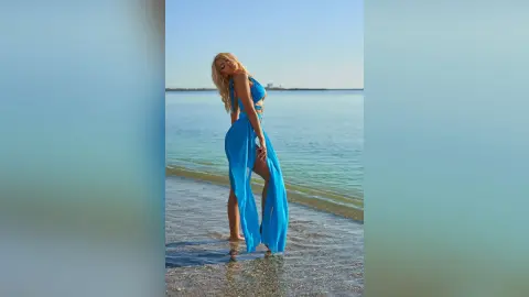 Anastasia stands in shallow ocean water wearing a long, sheer blue dress with cutouts on her sides.