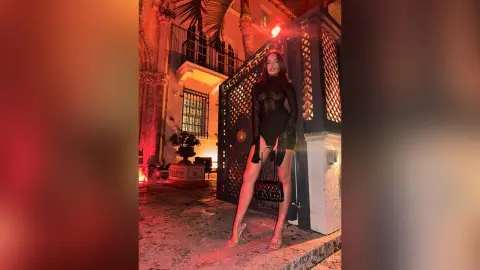 EvelynMills stands in a dark courtyard wearing a black long-sleeved mini dress and clear heels under a red glow.