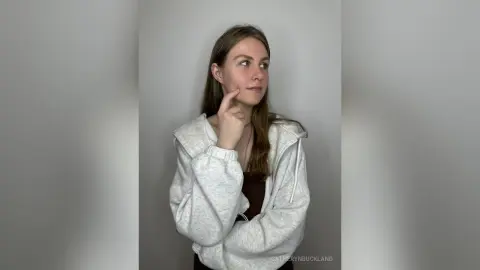 CatherynBuckland poses in a light grey hooded sweatshirt with her long brown hair down, touching her chin while looking to the side.