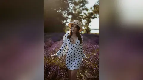 Dreamy portrait of a woman in a white polka dot dress and straw hat standing in a blooming lavender field.
