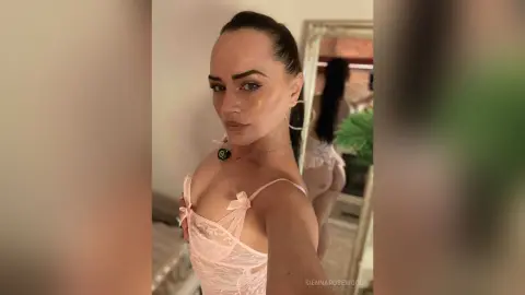 Young woman in pink lace lingerie posing for a selfie in a bright, modern bedroom setting.