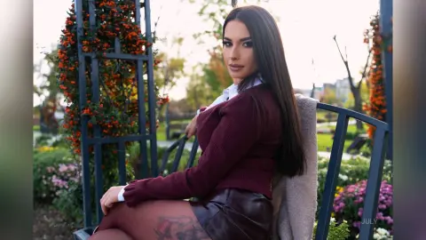Elegant woman in a burgundy sweater and leather skirt sitting on a garden bench during a sunny afternoon.