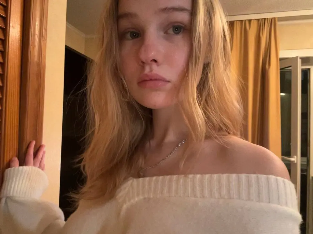 Blonde woman in a white off-the-shoulder sweater posing in a warm, cozy indoor setting.