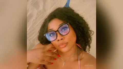 MandyJohn — MandyJohn wears blue sunglasses and lays on the sand with her dark curly hair spread out.