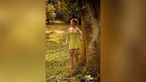Young woman in a lime green mini dress standing by a large tree in a sunlit forest setting.