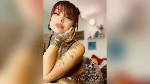 Tattooed woman in a black tank top posing with a dreamy expression in a bright, sunlit bedroom.