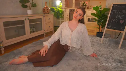 Young woman in a white lace blouse and brown skirt sitting on a rug in a cozy, sunlit living room.