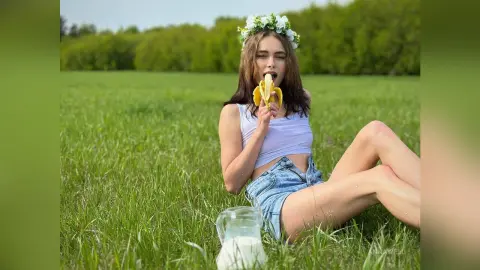 Young woman in white crop top and denim shorts eating a banana in a sunny green meadow.