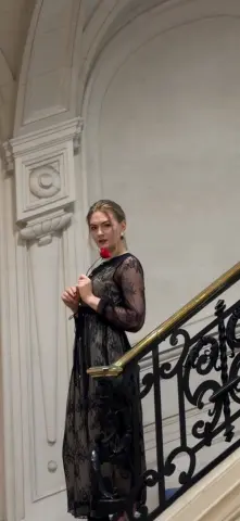 Elegant woman in a black lace dress holding a red rose on a classic white staircase.