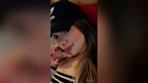Smiling young woman in a black Adidas cap and striped top relaxing in a cozy, dimly lit room.