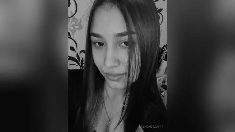 Close-up portrait of a young woman with long dark hair wearing a necklace in a moody black and white setting.