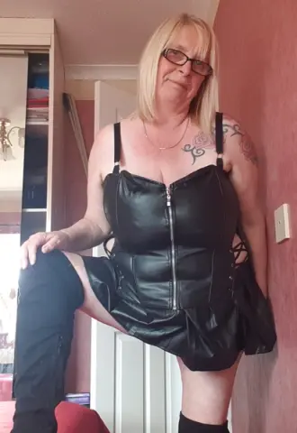 Blonde woman in black leather corset and shorts posing in a bedroom setting.