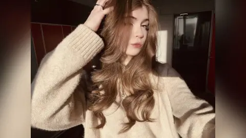 Young woman with long wavy hair wearing a cozy beige knit sweater in warm sunlight.