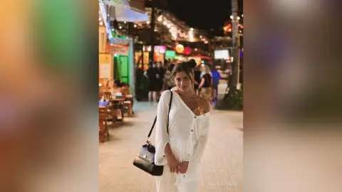 AnaMathers stands on a street at night wearing a white button-down shirt and white pants, carrying a black handbag over her shoulder.