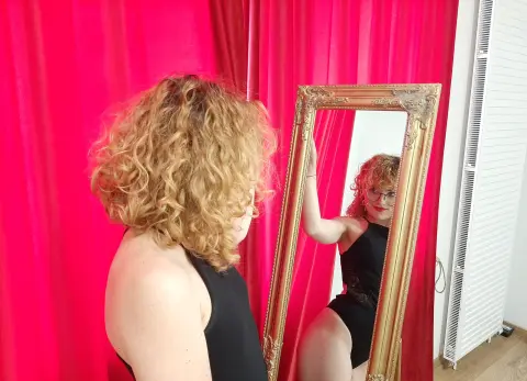 Woman with curly blonde hair in a black bodysuit looking at her reflection in a gold vintage mirror against red curtains.