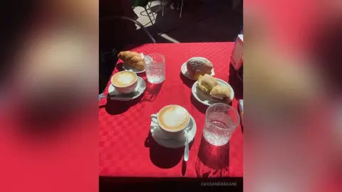 Breakfast spread with coffee, croissants, and water on a red tablecloth in bright morning sunlight.
