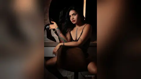 Moody portrait of a tattooed woman in a black lace bodysuit sitting in a dimly lit room.