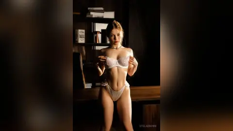 StelaBrown poses in white lace lingerie, pulling at her bra while standing in a dimly lit room.