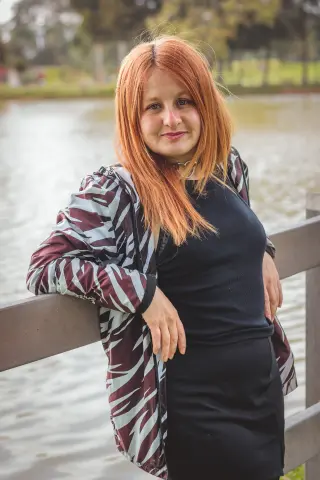 Red-haired woman in a black outfit and patterned jacket leaning on a wooden railing by a lake.